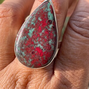 May include: A silver ring with a large, teardrop-shaped stone. The stone displays a striking pattern of red and teal hues. The ring is a statement piece, highlighting the natural beauty of the gemstone.