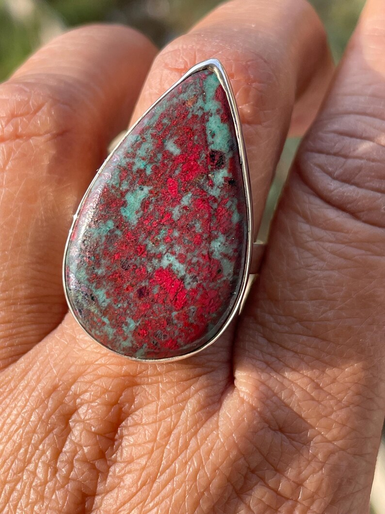 May include: A silver ring with a large, teardrop-shaped stone. The stone displays a striking pattern of red and teal hues. The ring is a statement piece, highlighting the natural beauty of the gemstone.