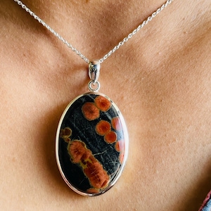 May include: Oval pendant necklace featuring a black stone with orange and red patterns, set in a silver-colored metal frame. The pendant hangs from a delicate silver chain, creating a striking contrast. A unique piece of jewelry.