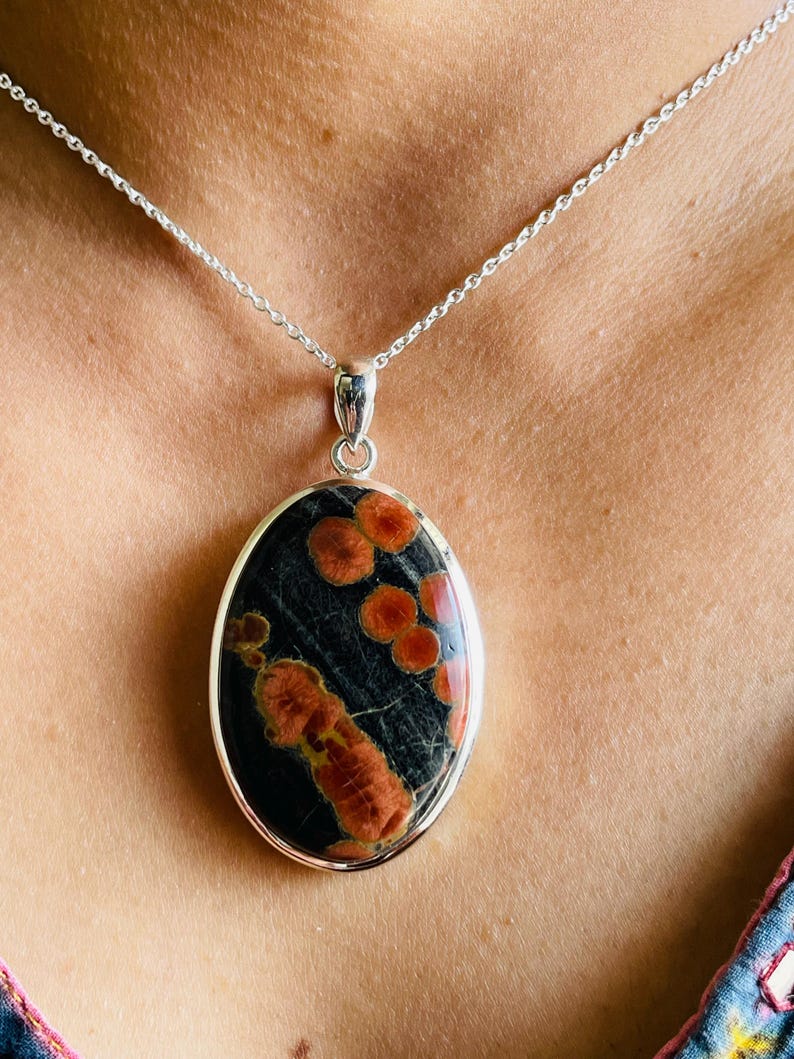 May include: Oval pendant necklace featuring a black stone with orange and red patterns, set in a silver-colored metal frame. The pendant hangs from a delicate silver chain, creating a striking contrast. A unique piece of jewelry.