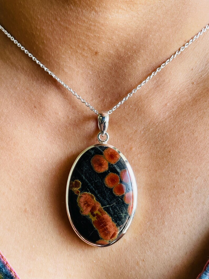May include: Oval pendant necklace featuring a dark stone with red and orange patterns, set in a silver-colored metal frame. The pendant hangs from a delicate silver chain, creating a striking contrast.