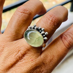 May include: A silver ring featuring a large, oval, pale green gemstone. The ring has a cluster of small, round silver beads on one side of the gemstone. The ring is on a finger.