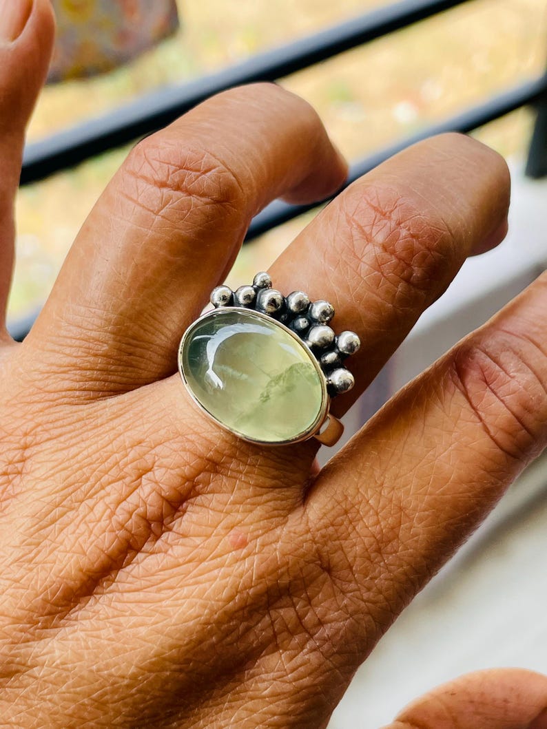 May include: A silver ring featuring a large, oval, pale green gemstone. The ring has a cluster of small, round silver beads on one side of the gemstone. The ring is on a finger.