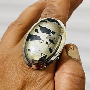 May include: A large oval ring featuring a polished metallic pyrite stone with distinct black inclusions set in a smooth silver band. The stone displays a reflective, brassy gold surface with irregular dark patterns across its face.