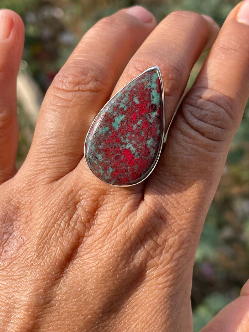 May include: A silver ring with a large, teardrop-shaped stone. The stone displays a mottled pattern of red and green colours, set within a silver band. The ring is shown on a hand.