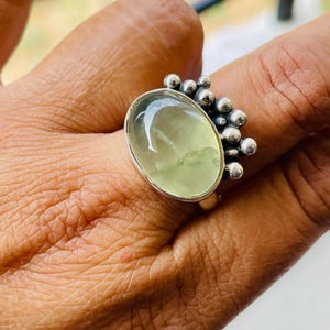 May include: A silver ring featuring a large, oval-shaped, pale green gemstone. The ring has a cluster of small, round silver beads on one side of the gemstone. The ring is set on a finger.