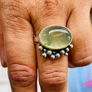 May include: A silver ring featuring a large, oval-shaped, pale green gemstone. The ring has a cluster of small, round silver beads on the band. The gemstone is set in a silver bezel.