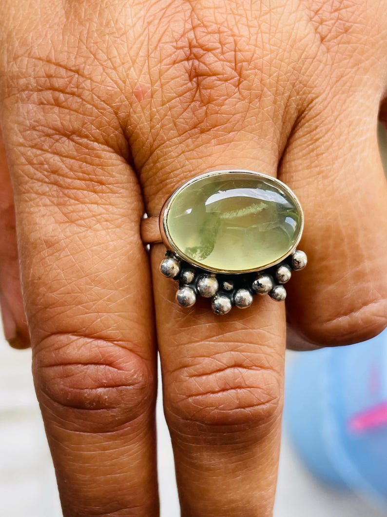May include: A silver ring featuring a large, oval-shaped, pale green gemstone. The ring has a cluster of small, round silver beads on the band. The gemstone is set in a silver bezel.