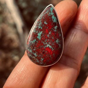 May include: A silver ring with a teardrop-shaped stone, showcasing a striking combination of red and teal colours. The stone's surface has a mottled appearance, with the red and teal hues creating a unique pattern. A bold piece of jewellery.