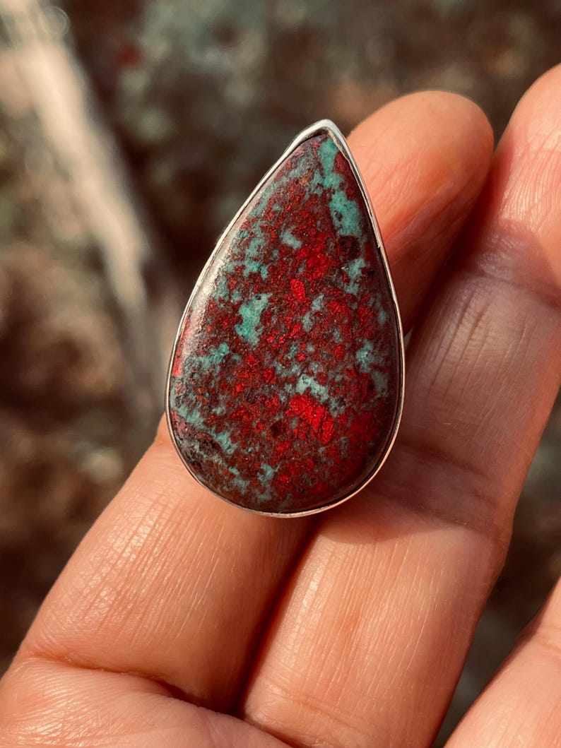 May include: A silver ring with a teardrop-shaped stone, showcasing a striking combination of red and teal colours. The stone's surface has a mottled appearance, with the red and teal hues creating a unique pattern. A bold piece of jewellery.