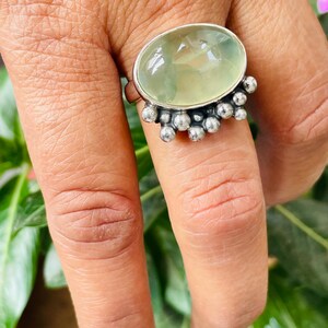 May include: A silver ring featuring a large, oval-shaped, pale green gemstone. The gemstone is surrounded by a silver setting with small, round silver accents. The ring is displayed on a hand, with green foliage in the background.