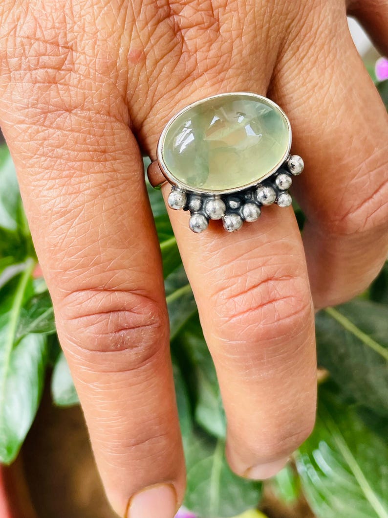 May include: A silver ring featuring a large, oval-shaped, pale green gemstone. The gemstone is surrounded by a silver setting with small, round silver accents. The ring is displayed on a hand, with green foliage in the background.