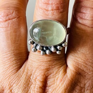 May include: Oval-shaped, light green gemstone ring with a silver band. The ring features a cluster of small silver beads around the gemstone. The ring is worn on a finger, showcasing the design and color of the jewelry.