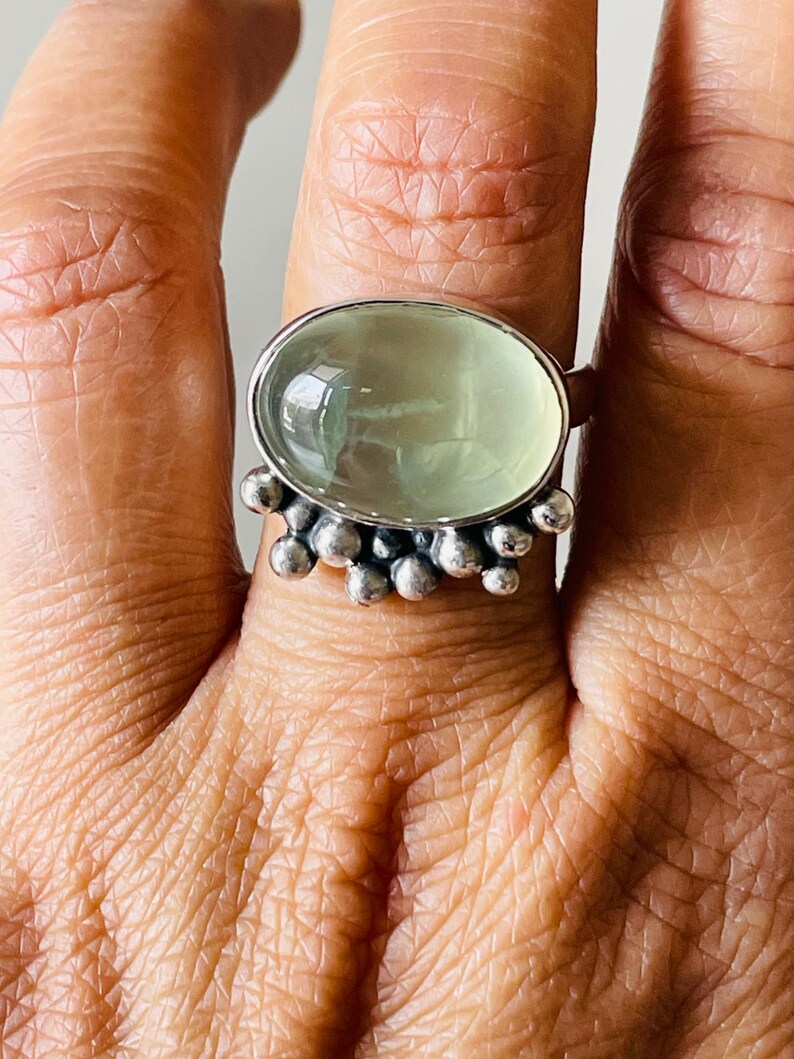 May include: Oval-shaped, light green gemstone ring with a silver band. The ring features a cluster of small silver beads around the gemstone. The ring is worn on a finger, showcasing the design and color of the jewelry.