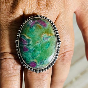 Könnte beinhalten: Ein ovaler Ring mit einem großen, polierten Cabochon-Stein mit einem gesprenkelten grünen und leuchtend magentafarbenen Muster. Der Stein ist in eine silberfarbene Metallfassung mit einem dekorativen Perlenrand eingefasst, der den Edelstein umrahmt.