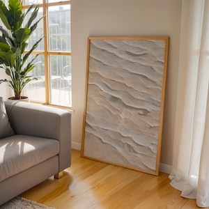 Puede incluir: Una obra de arte enmarcada con un diseño abstracto texturizado en tonos beige y crema. La obra se exhibe contra una pared neutra, junto a una ventana y un sofá gris. Una gran planta verde añade un elemento natural a la habitación.