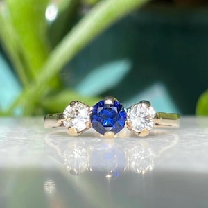 May include: A gold ring featuring a central, round, deep blue gemstone flanked by two smaller, clear, round gemstones. The ring is set against a blurred background of green foliage and a reflective surface.