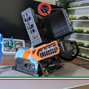 Puede incluir: Una caja de ordenador azul y naranja con varios componentes, incluyendo una placa base, una tarjeta gráfica y un refrigerador. Una figura azul está cerca. El fondo incluye una exhibición de modelos de coches.