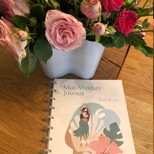 May include: A cream-colored journal with a spiral binding. The cover features a colorful illustration of a woman in a hula skirt dancing in front of a tropical flower and leaves. The text on the cover reads "Mut-Mindset Journal" and "Vive la vie".