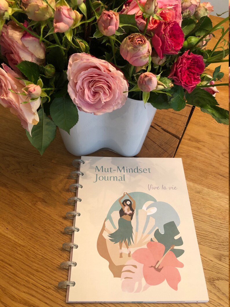 May include: A cream-colored journal with a spiral binding. The cover features a colorful illustration of a woman in a hula skirt dancing in front of a tropical flower and leaves. The text on the cover reads "Mut-Mindset Journal" and "Vive la vie".
