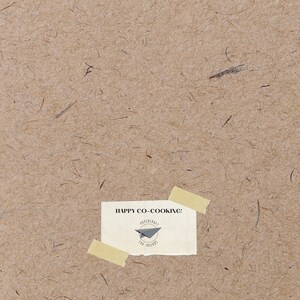 May include: A tan paper background with a yellow sticky note attached to it. The note says "HAPPY CO-COOKING!" and has a small black and white logo with a paper airplane and the text "PAPERCRAFT FOR FRIENDS".