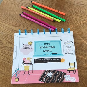 May include: A white spiral bound journal with a colorful illustration of a person sitting at a desk with a computer. The journal has the text "Mein Mindmapping Journal" on the cover. The text "Kreativität ist Intelligenz, die Spass hat. - Albert Einstein" is also on the cover.