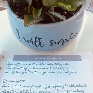 May include: A blue ceramic pot with the text "I will survive" on the side. The pot is filled with a succulent plant. The text "Dein Mutmacher-Journal" is written on a piece of paper below the pot. The paper has text about accepting responsibility and changing your life for the better.