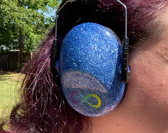 May include: Blue earmuffs with a glitter finish and a rainbow infinity symbol design. The earmuffs are worn by a person with purple hair.