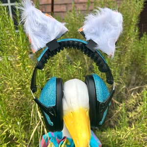 May include: A white goose wearing a pair of blue glitter earmuffs with a white furry headband with orange and black patterned ears.