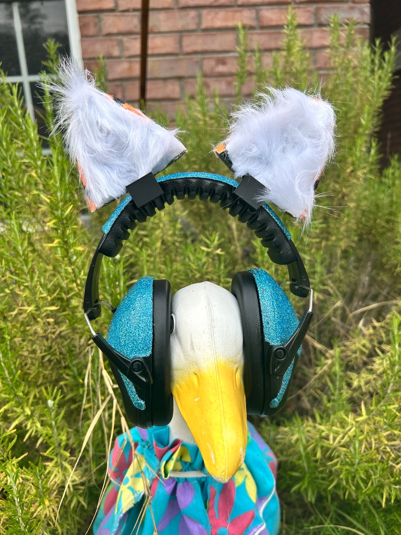 May include: A white goose wearing a pair of blue glitter earmuffs with a white furry headband with orange and black patterned ears.