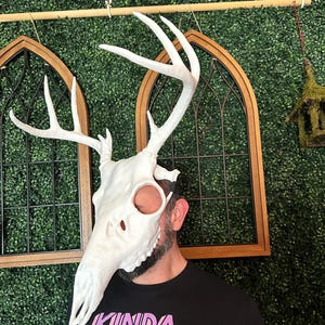May include: A white deer skull mask with large antlers. The mask covers the face and head, with eye and nose openings. The person wearing the mask is wearing a black t-shirt with the text "KINDA CHUNKY" in colorful letters.