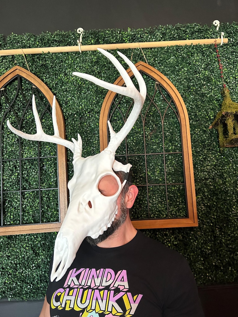 May include: A white deer skull mask with large antlers. The mask covers the face and head, with eye and nose openings. The person wearing the mask is wearing a black t-shirt with the text "KINDA CHUNKY" in colorful letters.