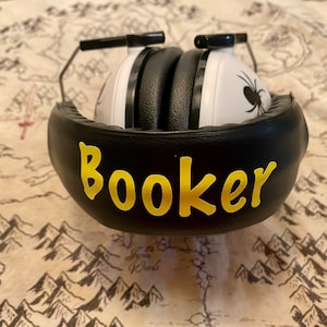 May include: Black noise-canceling headphones with a white earcup featuring a black spider design. The headphones have a black padded headband with the name "Booker" written in yellow letters.