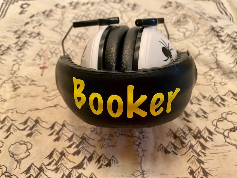 May include: Black noise-canceling headphones with a white earcup featuring a black spider design. The headphones have a black padded headband with the name "Booker" written in yellow letters.