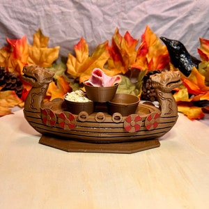 May include: A brown wooden Viking ship shaped serving tray with three small bowls. The bowls are filled with snacks, including pink and white candy and a white powder. The tray is sitting on a light brown wooden surface.