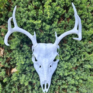 May include: A white deer skull with large antlers, set against a backdrop of green foliage. The skull has detailed features and a textured surface. The antlers are long and curved, adding to the skull's striking appearance. A decorative piece for home decor.