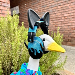 May include: A white goose wearing a blue and black pair of headphones with cat ears, and a blue, pink, and yellow floral print shirt.