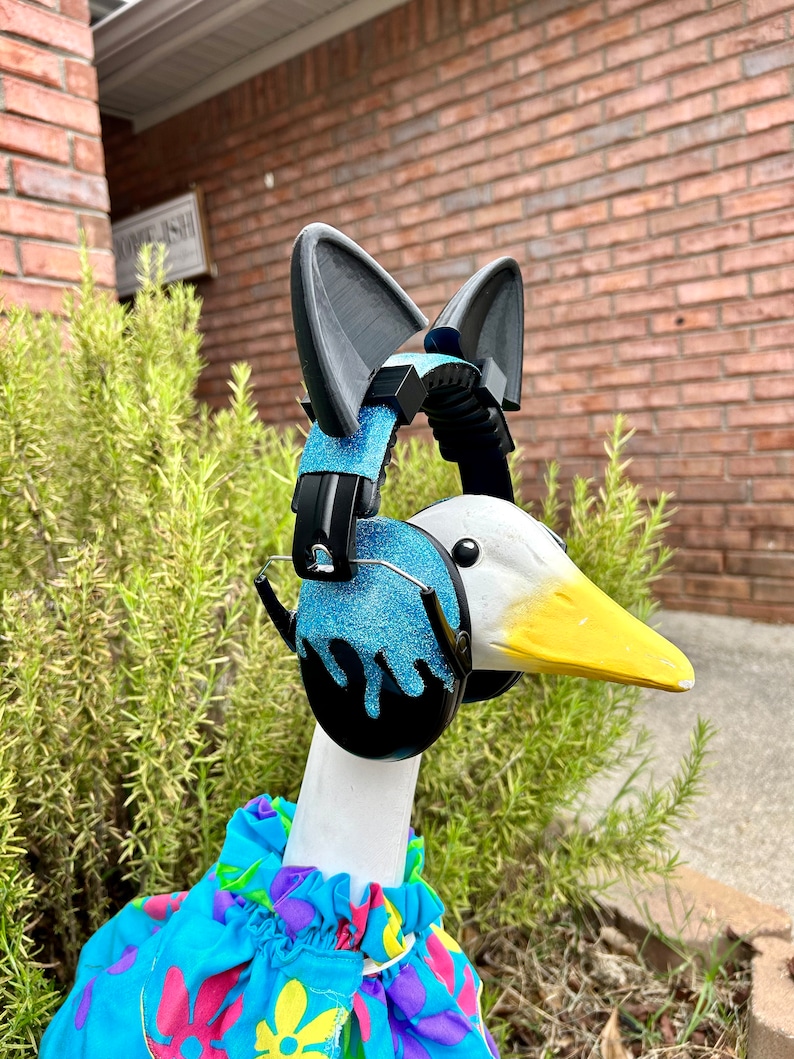 May include: A white goose wearing a blue and black pair of headphones with cat ears, and a blue, pink, and yellow floral print shirt.