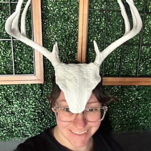 May include: A white deer antler headpiece with a detailed skull design. The antlers are long and curved, with multiple points. The headpiece is worn by a person. The background is a green wall with brown framed windows.