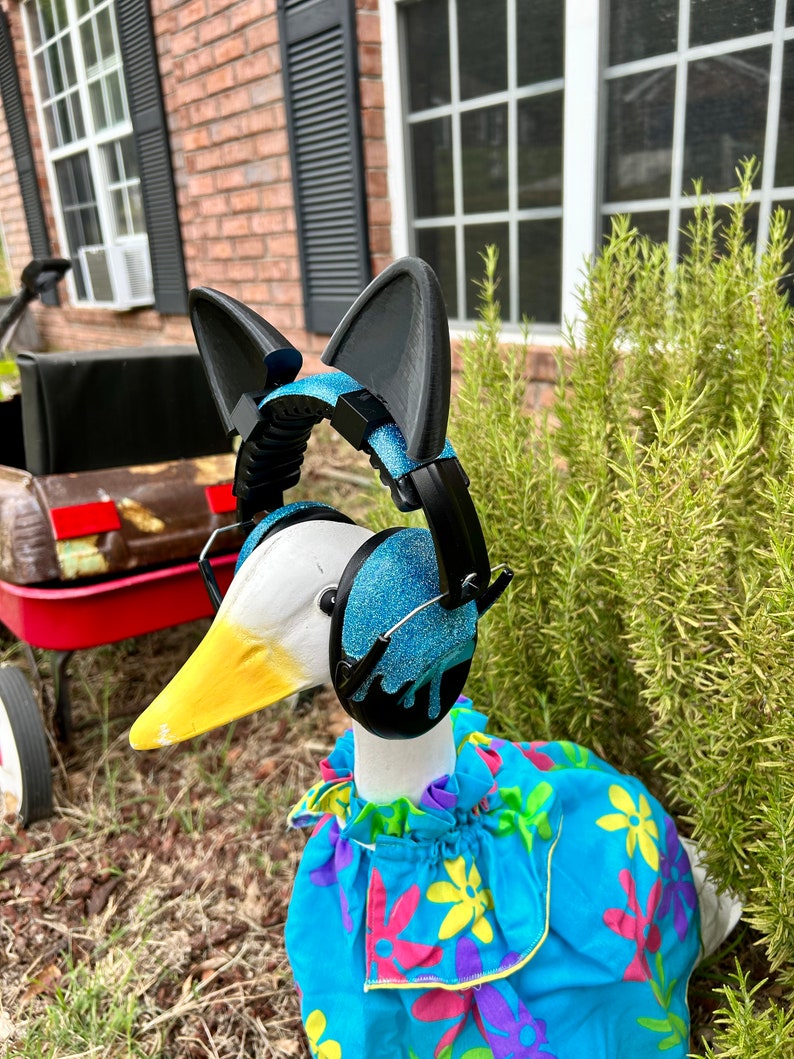 May include: A white goose wearing a blue and black pair of headphones with cat ears, a blue floral dress, and a yellow beak. The goose is standing on a bed of brown mulch.