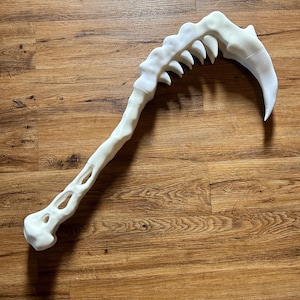 May include: A white, bone-like prop weapon featuring a long, curved handle with open, hollow sections and a jagged, tooth-lined blade at the top. The object has a textured, matte finish and resembles a prehistoric or fantasy-inspired scythe.
