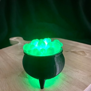 May include: A black cauldron with a green glow, filled with glowing green balls. The cauldron is sitting on a wooden surface.