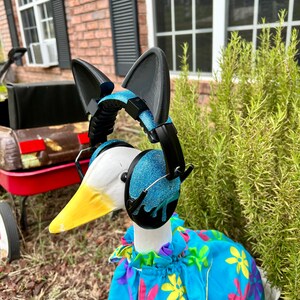 May include: A white goose wearing a blue and black pair of headphones with cat ears, a blue floral dress, and a yellow beak. The goose is standing on a bed of brown mulch.