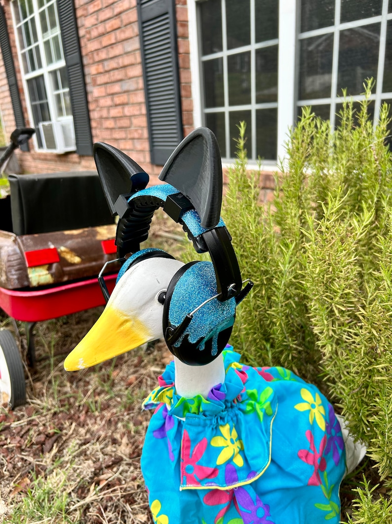 May include: A white goose wearing a blue and black pair of headphones with cat ears, a blue floral dress, and a yellow beak. The goose is standing on a bed of brown mulch.