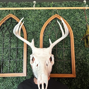 May include: A white deer skull mask with large antlers. The mask covers the face of a person wearing a black t-shirt with the text "KINDA CHUNKY" in pink and yellow. The background includes decorative wooden framed windows and a green hedge.