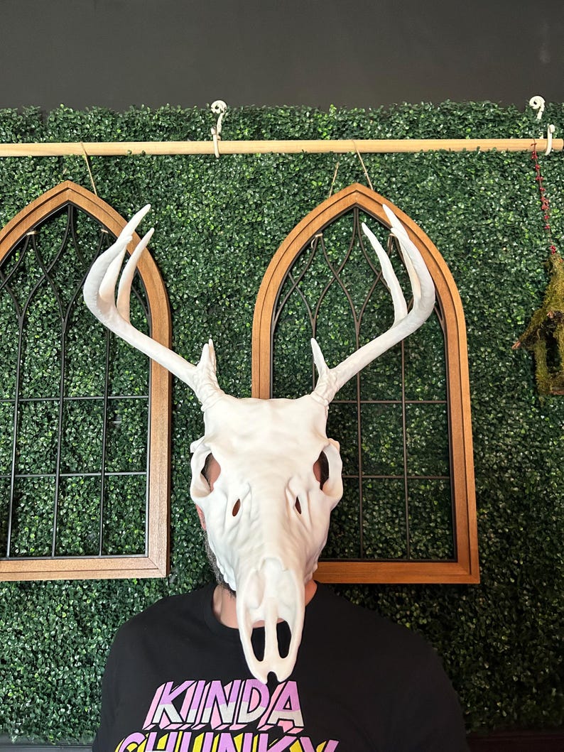 May include: A white deer skull mask with large antlers. The mask covers the face of a person wearing a black t-shirt with the text "KINDA CHUNKY" in pink and yellow. The background includes decorative wooden framed windows and a green hedge.