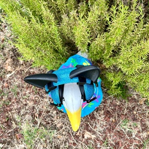 May include: A white goose wearing a blue and pink patterned shirt with black earmuffs. The goose is standing on a bed of brown mulch and green grass.
