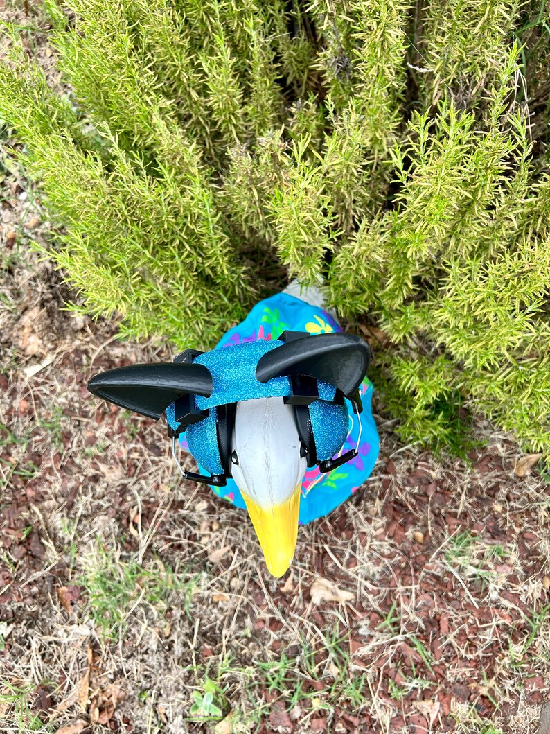 May include: A white goose wearing a blue and pink patterned shirt with black earmuffs. The goose is standing on a bed of brown mulch and green grass.