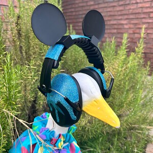 May include: A white goose wearing blue glitter headphones with black Mickey Mouse ears. The goose is wearing a blue, pink, and yellow floral shirt.