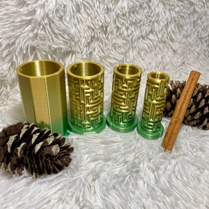 May include: Four gold and green candle holders with a maze pattern. The candle holders are arranged in a row from largest to smallest. There are two pine cones and a cinnamon stick in the background.